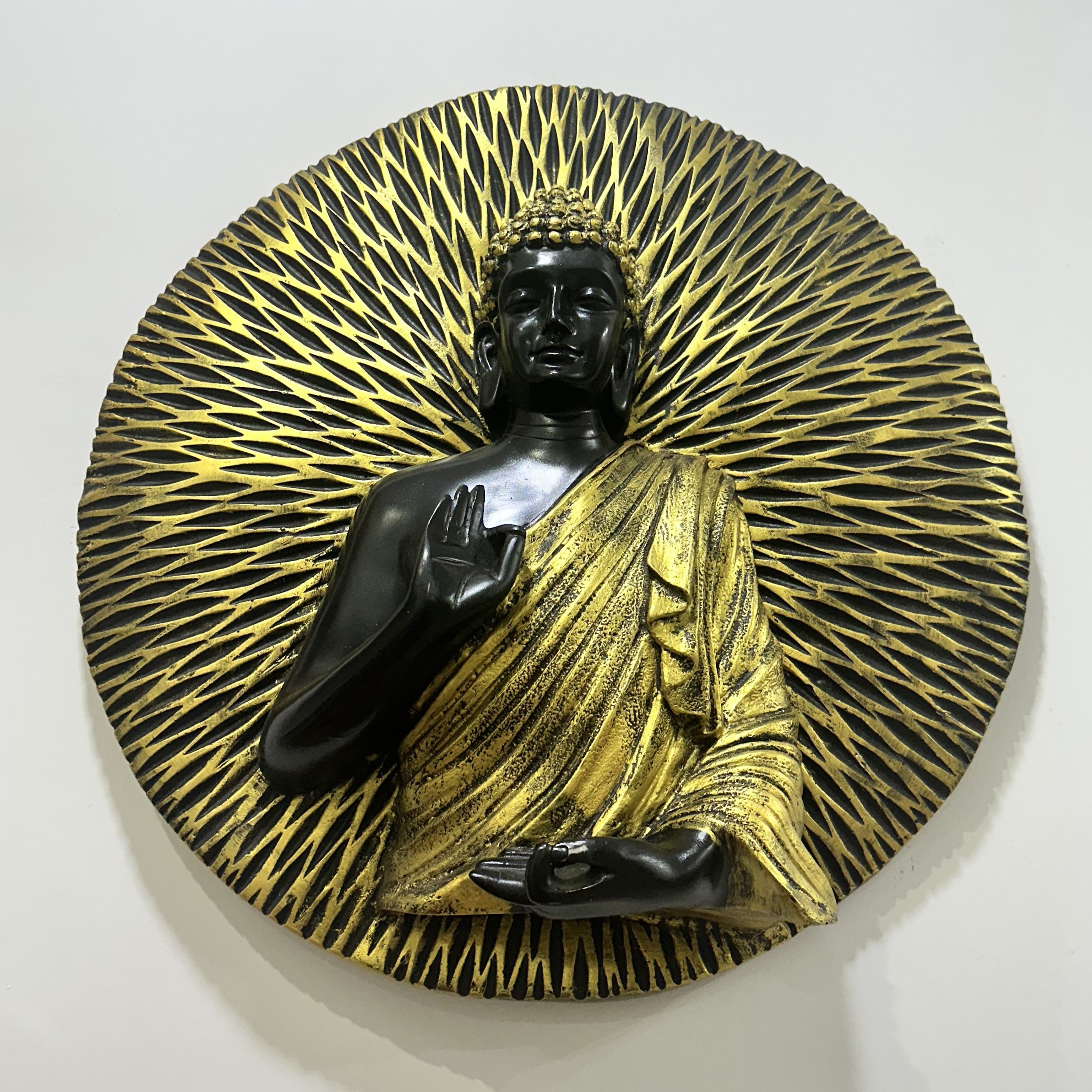 A circular Buddha wall sculpture featuring a black figure with a gold halo and robe, mounted on a light beige wall.
