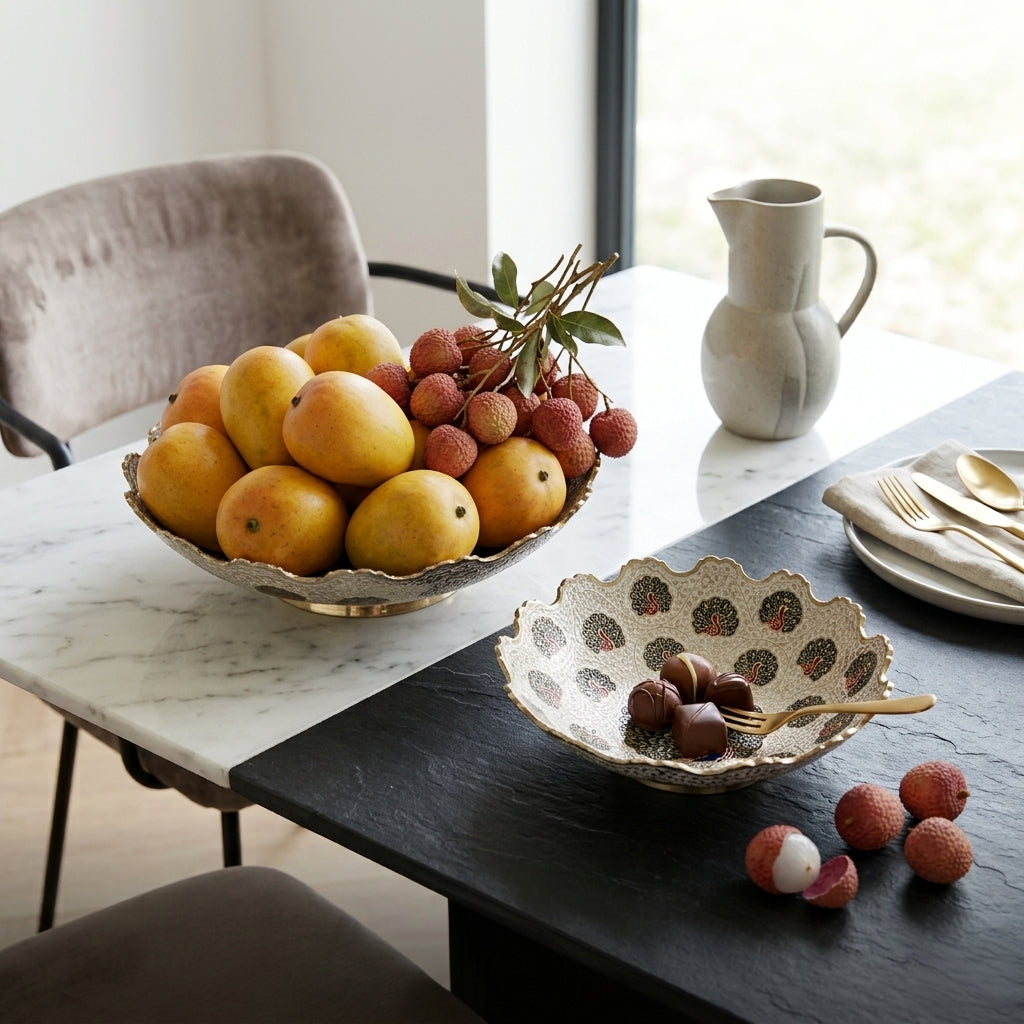 A styled dining table scene with a bowl of lychee fruit and pears, a small decorative bowl with chocolate-covered nuts and a fork, a ceramic pitcher, plates with gold-toned cutlery, and chairs in a bright indoor setting.