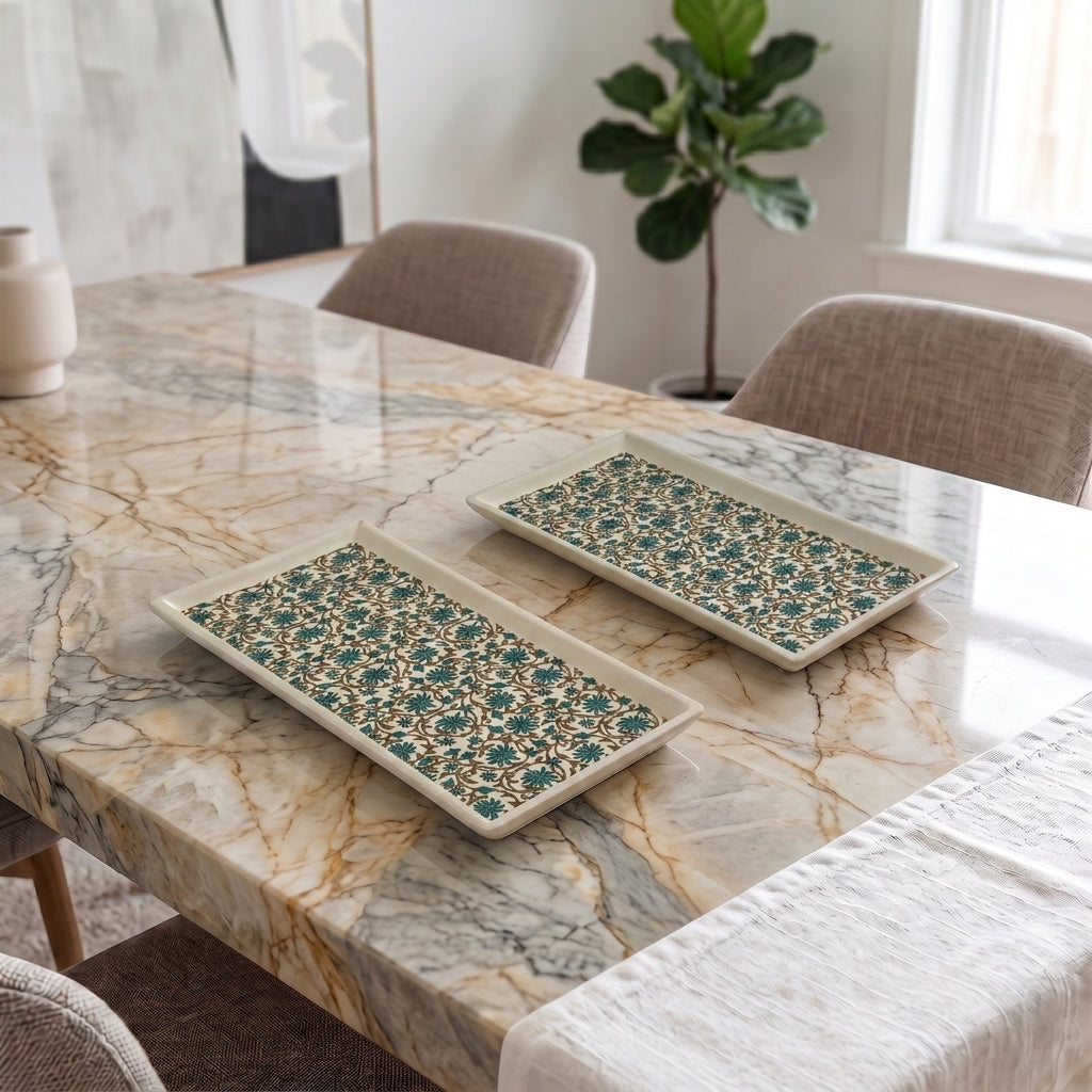 Two rectangular ceramic serving trays with a teal and beige floral pattern, displayed on a marble dining table in a bright, modern room.